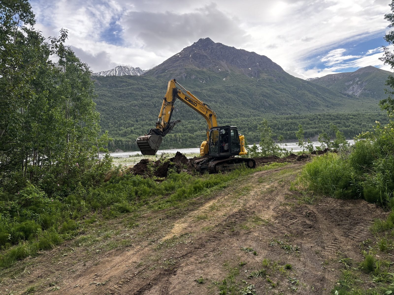 Excavator performing earthwork and site grading on a riverfront lot with an Alaska mountain peak and river behind — Mat-Su Valley