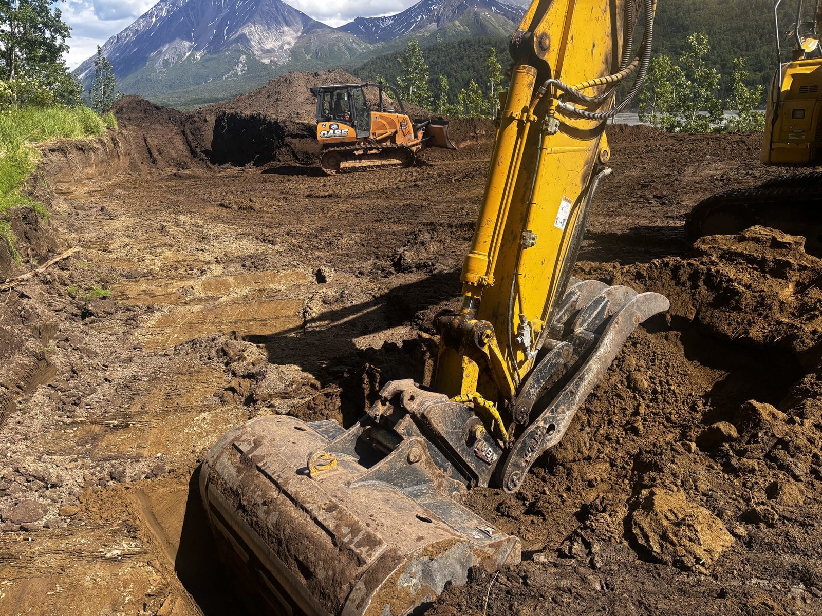 Excavator digging a trench during site excavation with a second machine and Alaska mountains in the background — Mat-Su Valley