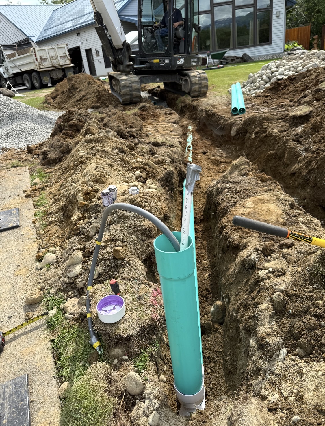 Septic system installation — deep trench with green PVC pipes being set next to a residential home