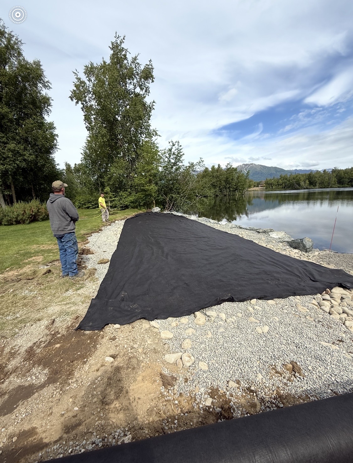 Crew installing geotextile erosion-control fabric along a lakeshore riprap wall, Alaska mountains in background