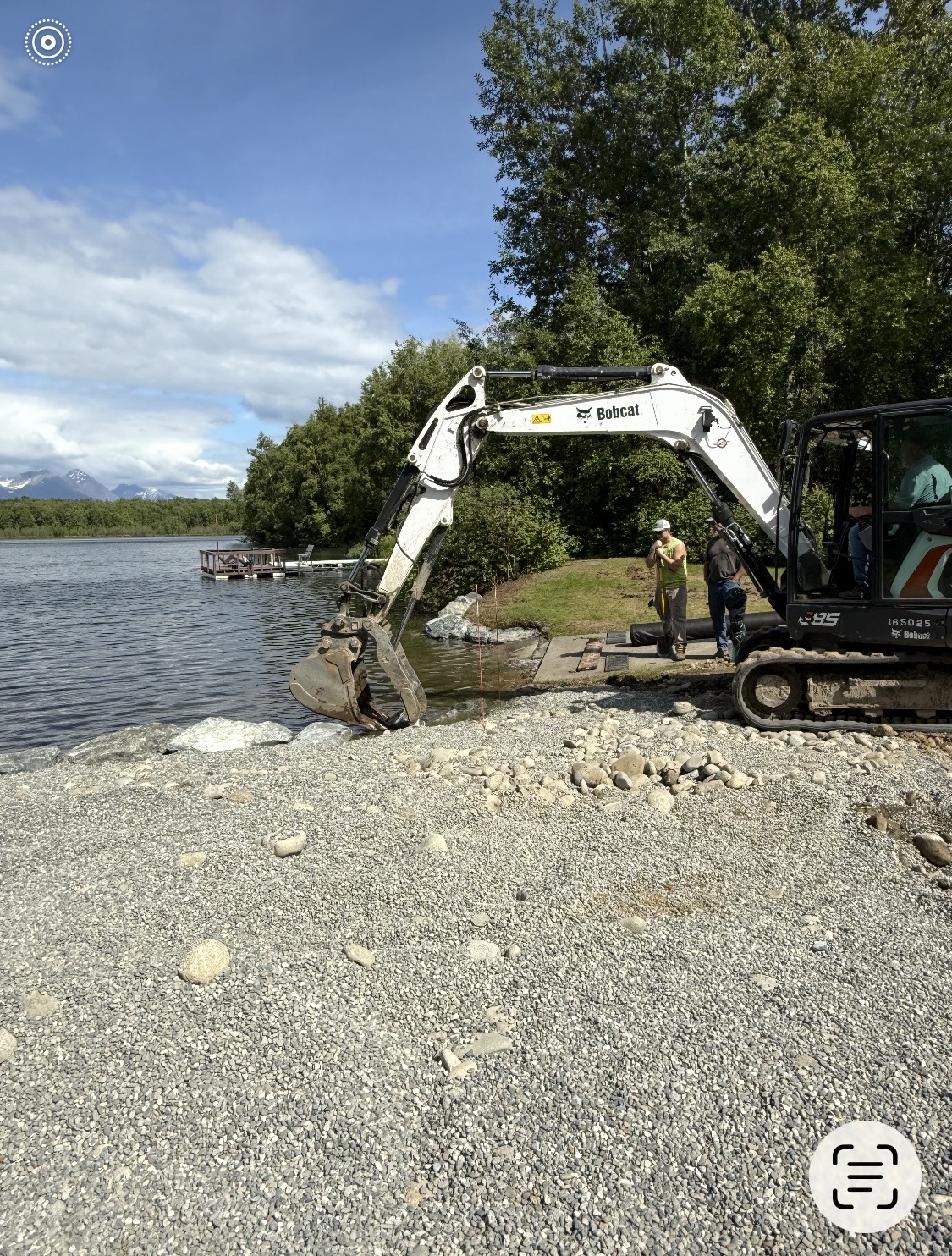 Excavator placing large riprap rocks along a lakeshore with worker directing placement