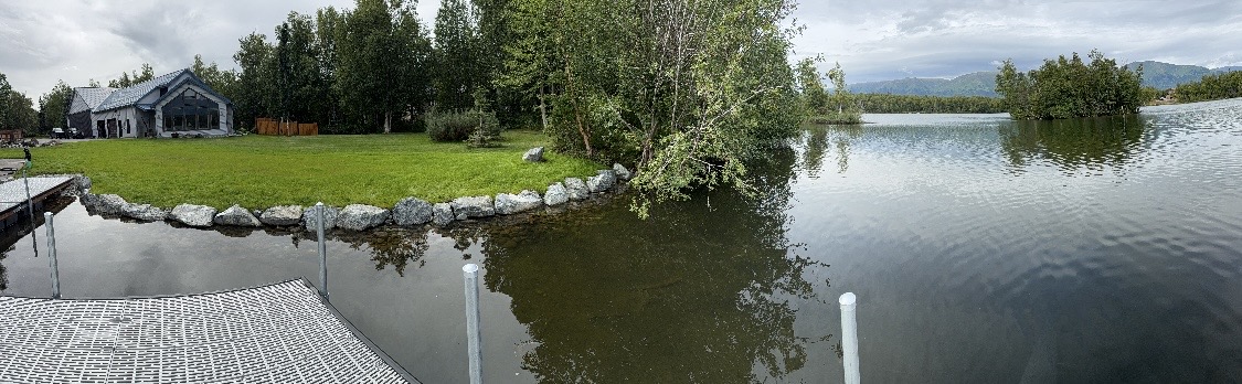 Panoramic view from dock of completed shoreline stabilization project with rock wall, lawn, and house — Mat-Su Valley